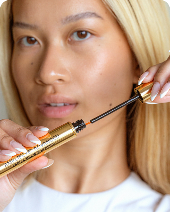 A person with blonde hair holds a gold tube of Amplifeye® Advanced Lash & Brow Fortifier, with the applicator brush extended.
