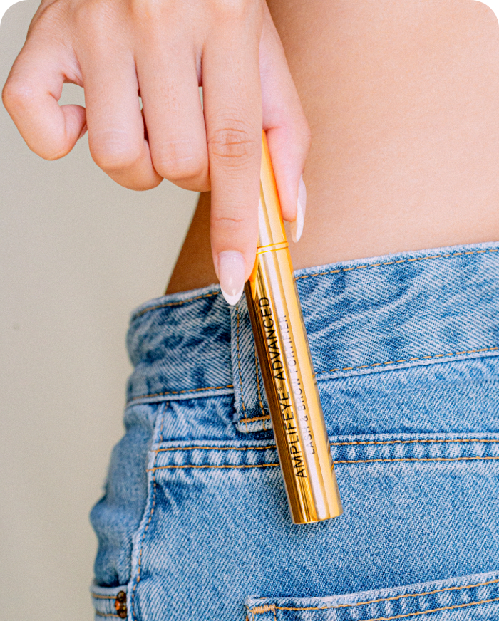 A hand with manicured nails holds a gold tube of Amplifeye® Advanced Lash & Brow Fortifier, with the product label clearly visible, positioned against a person's torso wearing blue jeans.