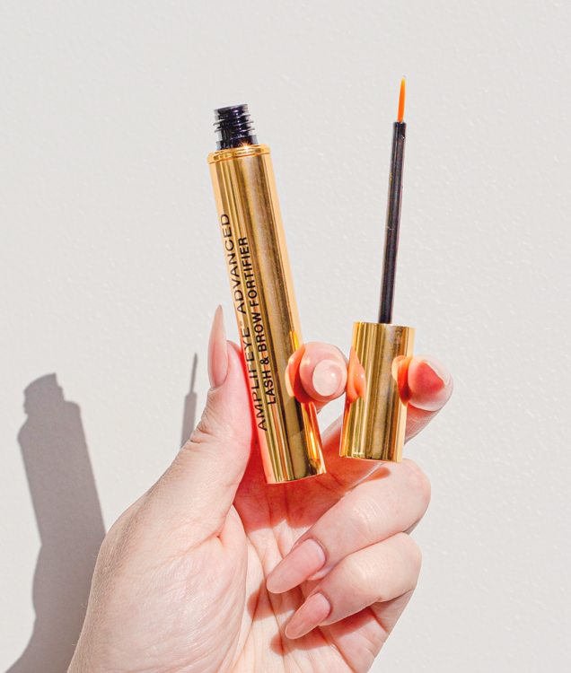 A hand holds an open gold tube of Amplifeye® Advanced Lash & Brow Fortifier, with the cap and applicator brush next to it.