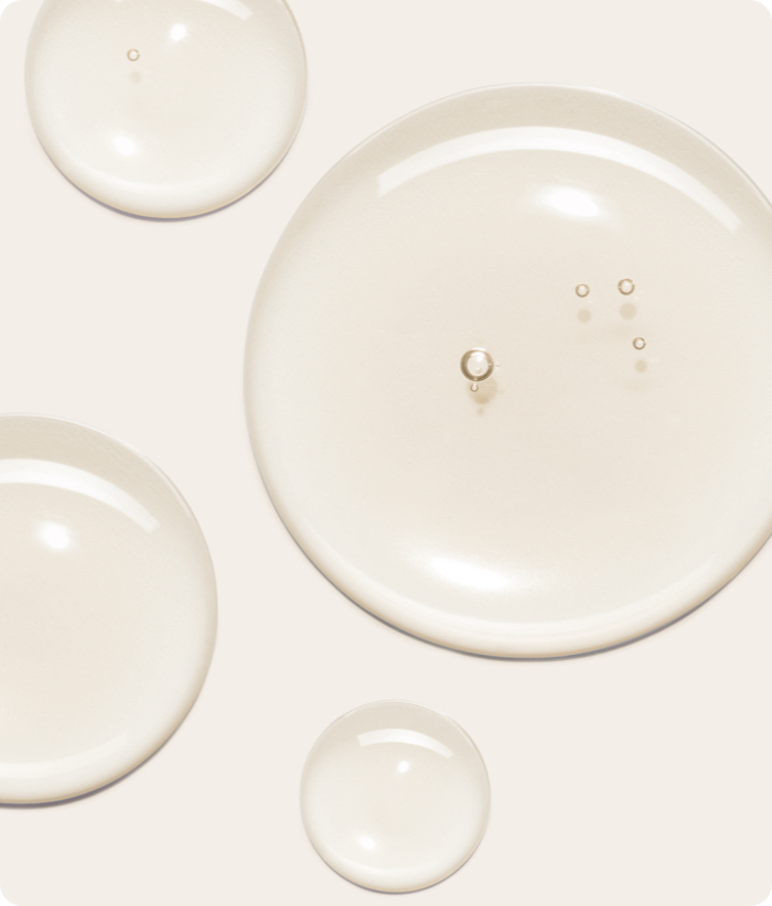 A close-up macro shot of four clear, gel-like droplets with a subtle yellowish tint on a light background. One large droplet has small air bubbles in it.