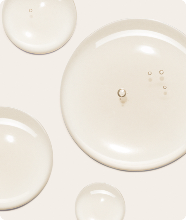 A close-up macro shot of four clear, gel-like droplets with a subtle yellowish tint on a light background. One large droplet has small air bubbles in it.